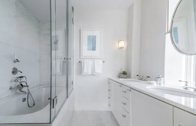 A white bathroom with a tub enclosed by a glass shower screen, a double vanity with a round mirror, and towels on a wall rail.