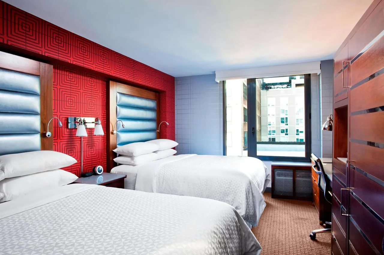 A bedroom with two double beds, a red patterned wall, a wooden wardrobe, a desk, and a window showing neighbouring buildings.