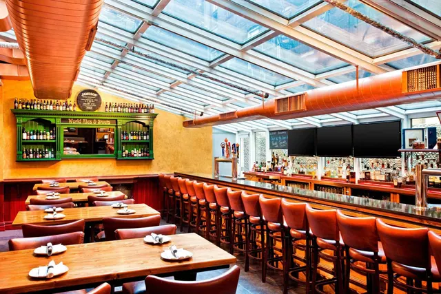 A skylit bar and dining room with leather bar stools, wooden tables set with plates and napkins, and shelves of bottles behind the bar.