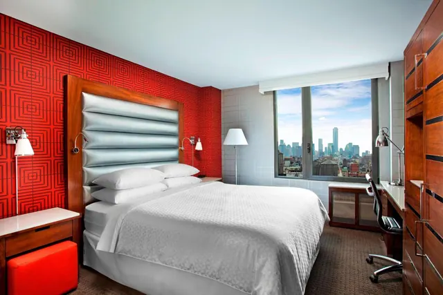 A bedroom with a king bed, a red patterned accent wall, a work desk, and a window looking out over a city skyline.