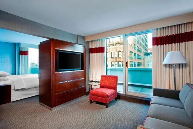 A hotel suite living area with a wall-mounted television in a wooden cabinet, a red armchair and a grey sofa, with a bed visible through the opening and sliding doors onto a balcony overlooking city buildings.
