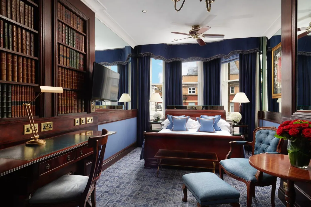 A bedroom with a double bed, built-in bookshelves, a writing desk and blue velvet curtains, with windows looking onto neighbouring buildings.