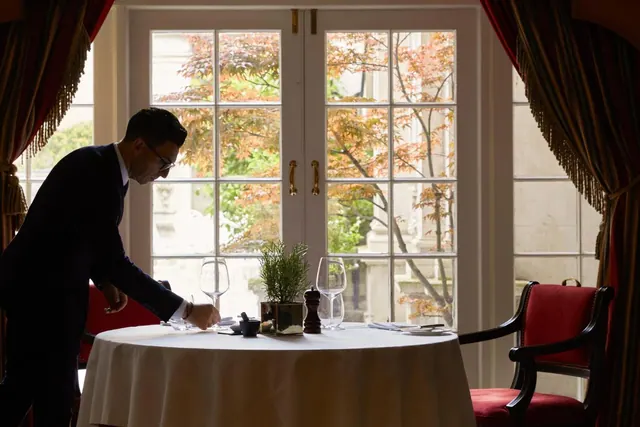 A waiter in a dark suit sets a round table with a white cloth, wine glasses and a potted herb in front of tall windows looking onto autumn trees and a stone building.