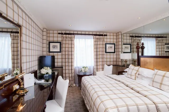 A hotel bedroom with a plaid-patterned double bed, a dark wood desk with a television and telephone, white chairs, and tall mirrors on the right wall.