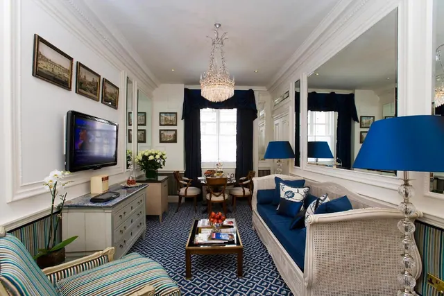 A living area in a hotel suite with a blue cane sofa, a wall-mounted television, a chandelier and a dining table visible beyond.