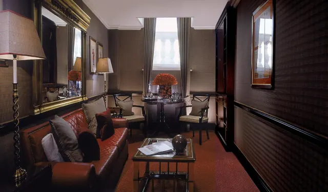 A living room with a red leather sofa, two armchairs, a round side table by a curtained window, and a tall dark wooden cabinet on the right.