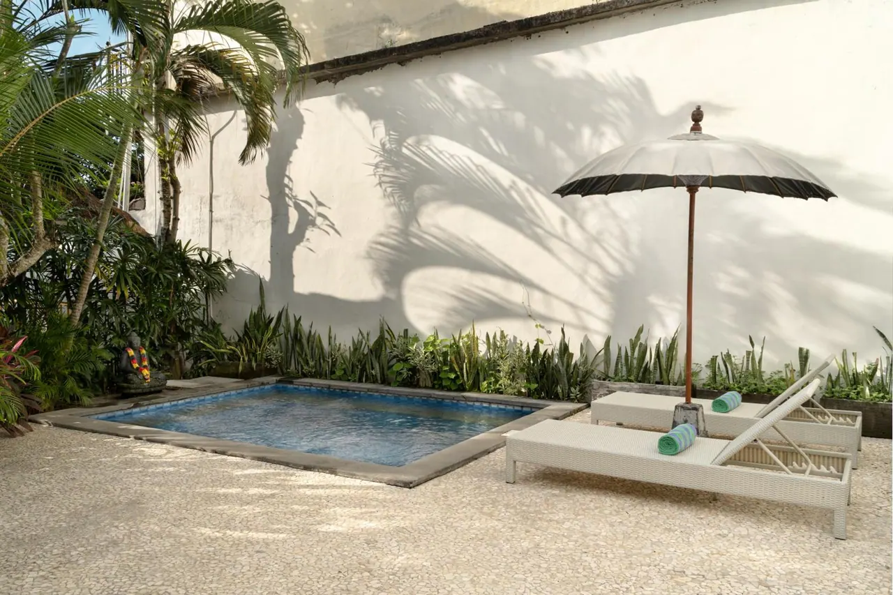 A small plunge pool in a walled courtyard, with two white sun loungers, teal towels and a black parasol beside it.