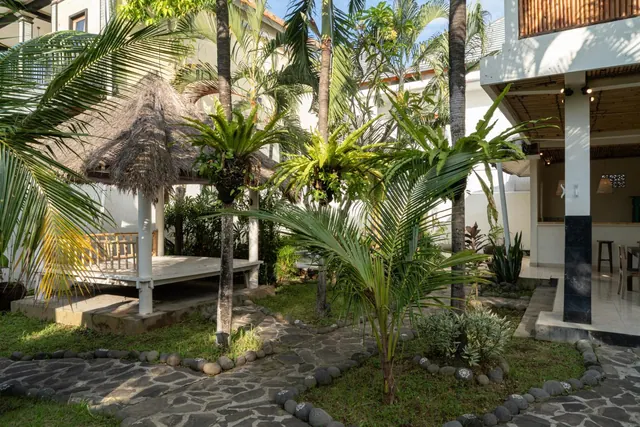 A landscaped garden courtyard with palm trees, a thatched-roof pavilion and a river-stone pathway, beside a building with a bar counter and stools.