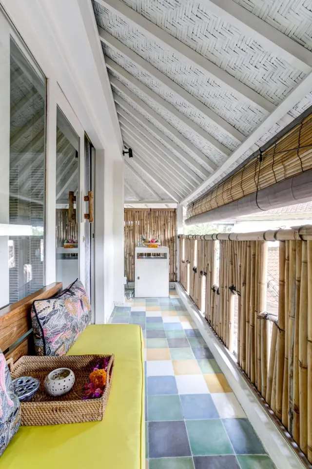 A covered veranda with a lime green cushioned daybed, patterned cushions, a rattan tray with a candle holder and flowers, bamboo screens along the side, and a checkered tile floor.