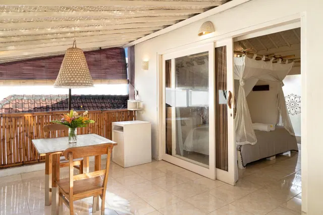 A covered terrace with a wooden dining table, two chairs and a bamboo railing, with glass doors open to a bedroom containing a canopy bed with a mosquito net.