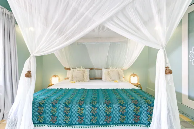 A four-poster bed with white sheer netting and a teal patterned throw sits between two wicker lamps in a mint-walled bedroom.