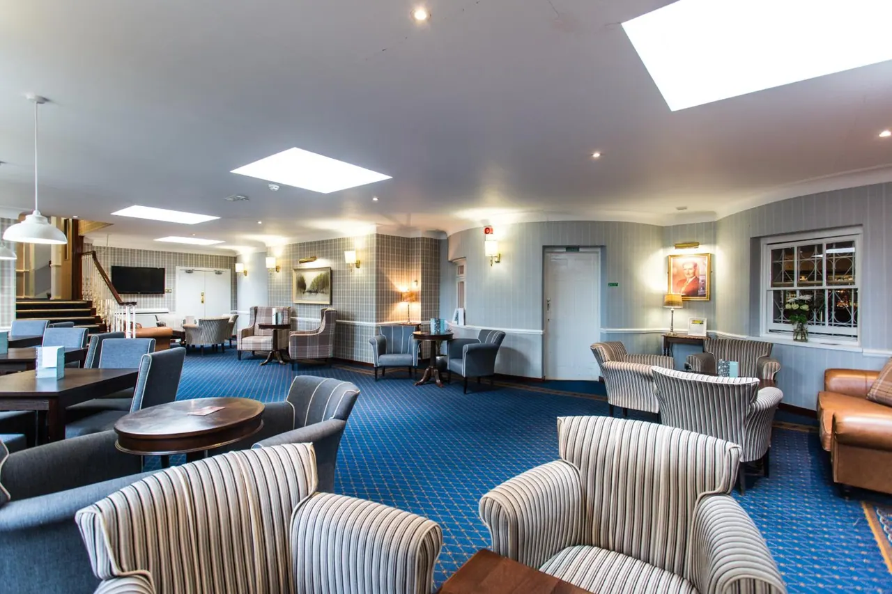 A hotel lounge with striped armchairs, wooden tables, blue patterned carpet, skylights and a wall-mounted television near the staircase.