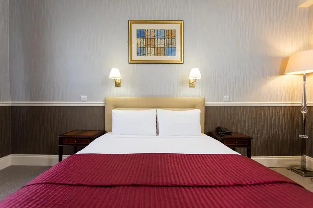 A double bed with white pillows and a red throw sits between two wooden bedside tables, with wall lamps and a framed picture above the headboard.