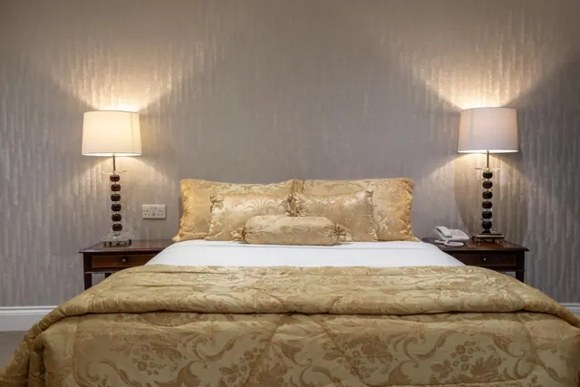 A double bed with gold patterned bedding and cushions sits between two lit bedside lamps against a textured grey wall.