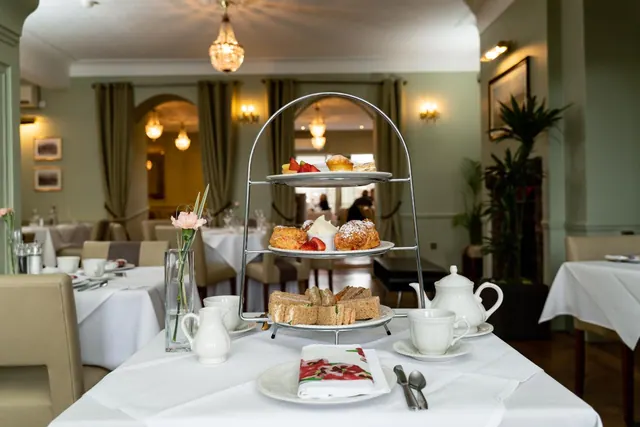 A three-tier afternoon tea stand with sandwiches, scones and cakes sits on a white-clothed table in a dining room with green walls and curtains.