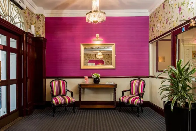 A lobby with a magenta wall, a crystal chandelier, two striped armchairs flanking a wooden console table, and a potted plant beside a glass partition.