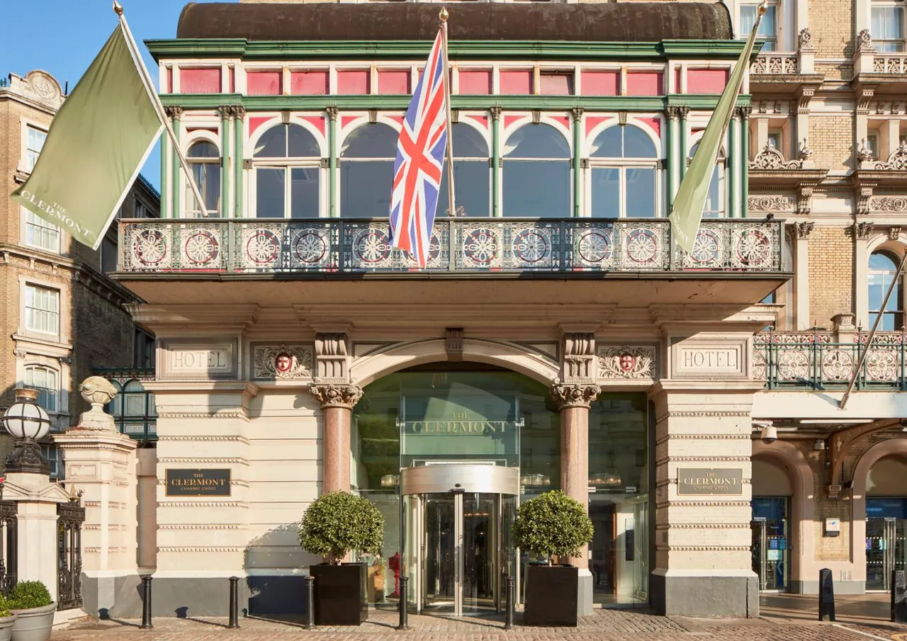 The Victorian street facade of The Clermont Charing Cross hotel, with a Union Jack and green flags, a revolving entrance door and clipped topiary planters at the entrance.