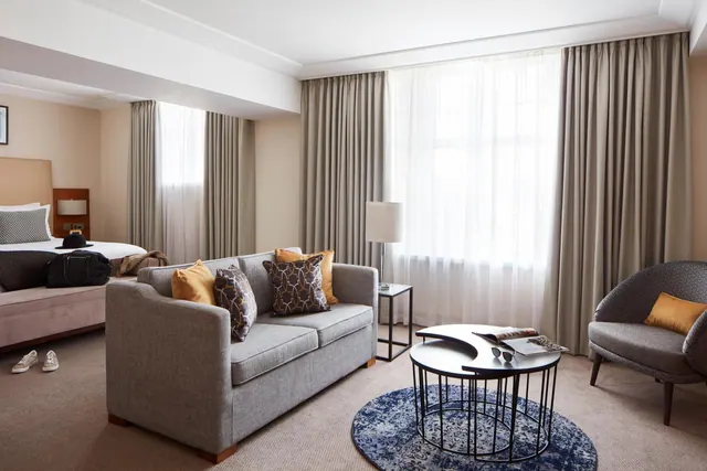 A hotel suite with a double bed on the left, a grey sofa and armchair, a round coffee table and a blue rug in front of tall windows with sheer curtains.