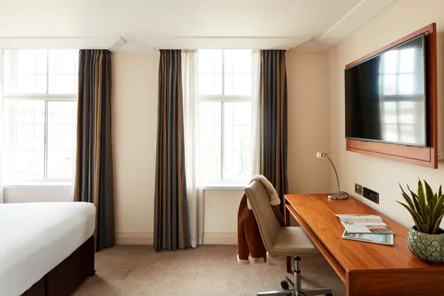 A hotel bedroom with a made bed on the left, striped curtains at two windows, a wooden desk with a chair and magazines, and a wall-mounted television.