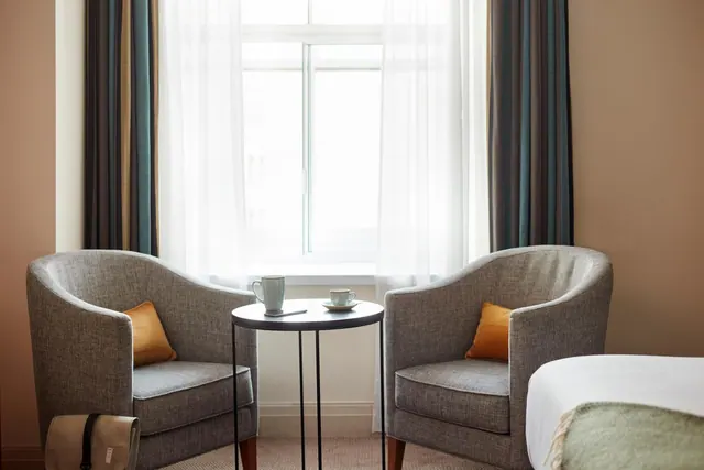 Two grey armchairs with orange cushions sit either side of a small round table by a window, with the corner of a bed visible at right.
