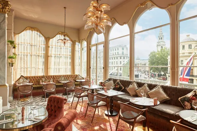A restaurant dining room with arched windows, banquette seating and set tables, looking out onto a church spire and historic buildings with a Union Jack flag.