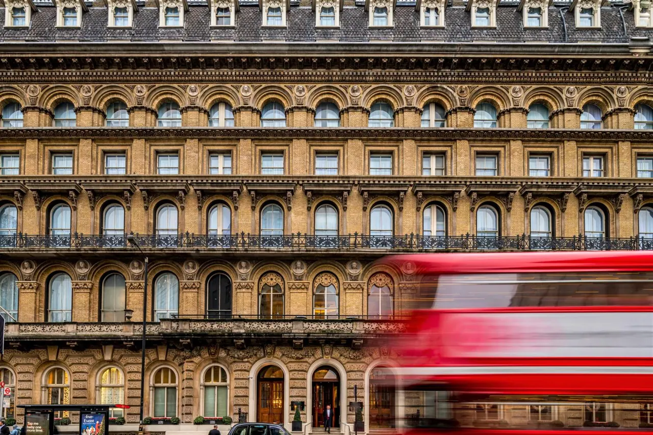 The facade of an ornate stone Victorian building with arched windows and a motion-blurred red double-decker bus passing in front.