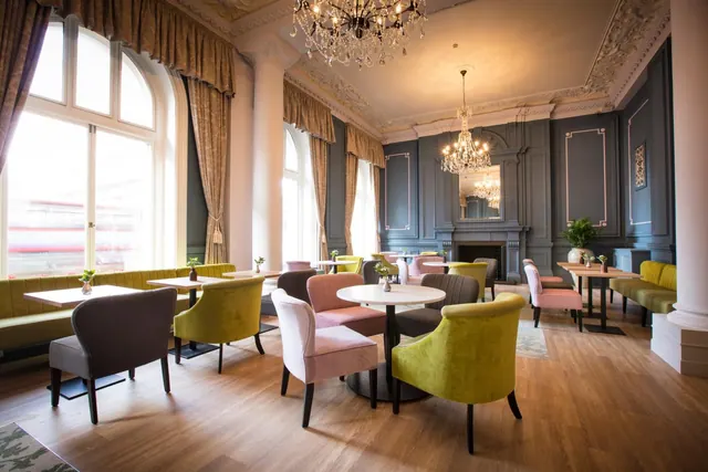 A lounge with tall arched windows, velvet armchairs and banquette seating, round marble-topped tables, and two chandeliers under an ornate ceiling.