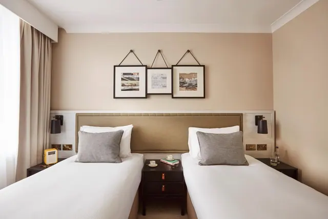 Two single beds with white bedding and grey cushions sit either side of a dark nightstand in a bedroom with beige walls and framed prints above the headboard.