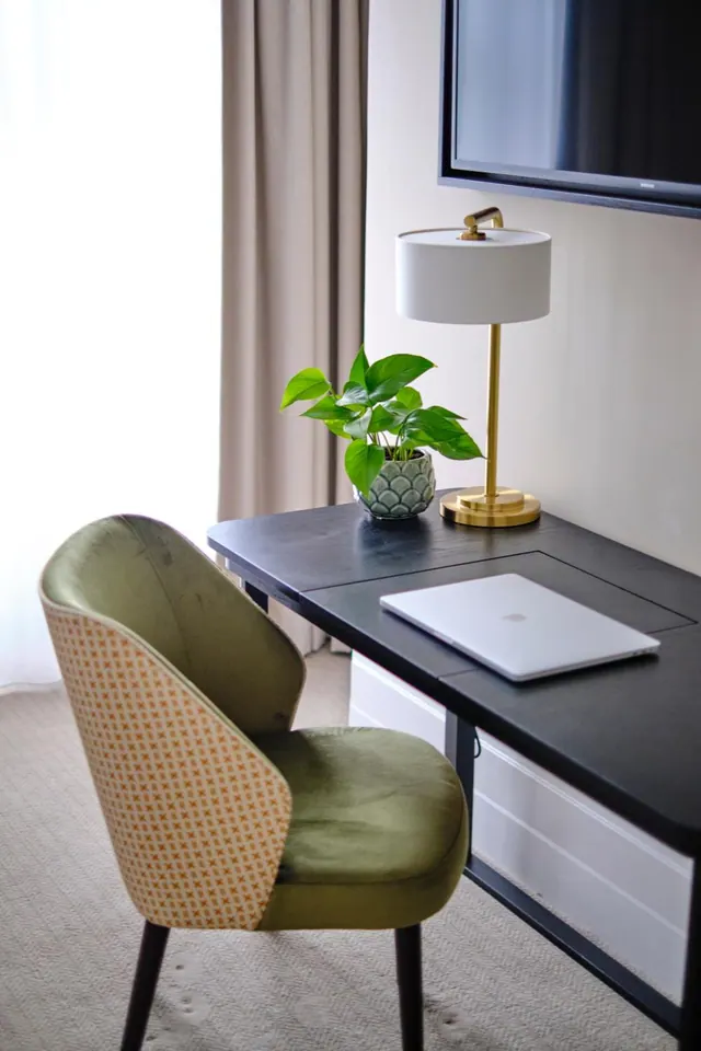 A black desk holds a brass table lamp, a potted plant and a closed silver laptop, with a green upholstered chair in front and a wall-mounted television above.