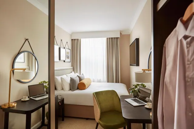 A double bed with cushions sits beside a window with sheer curtains, with two desks holding laptops, a wall-mounted television and a wardrobe with a hanging shirt in view.