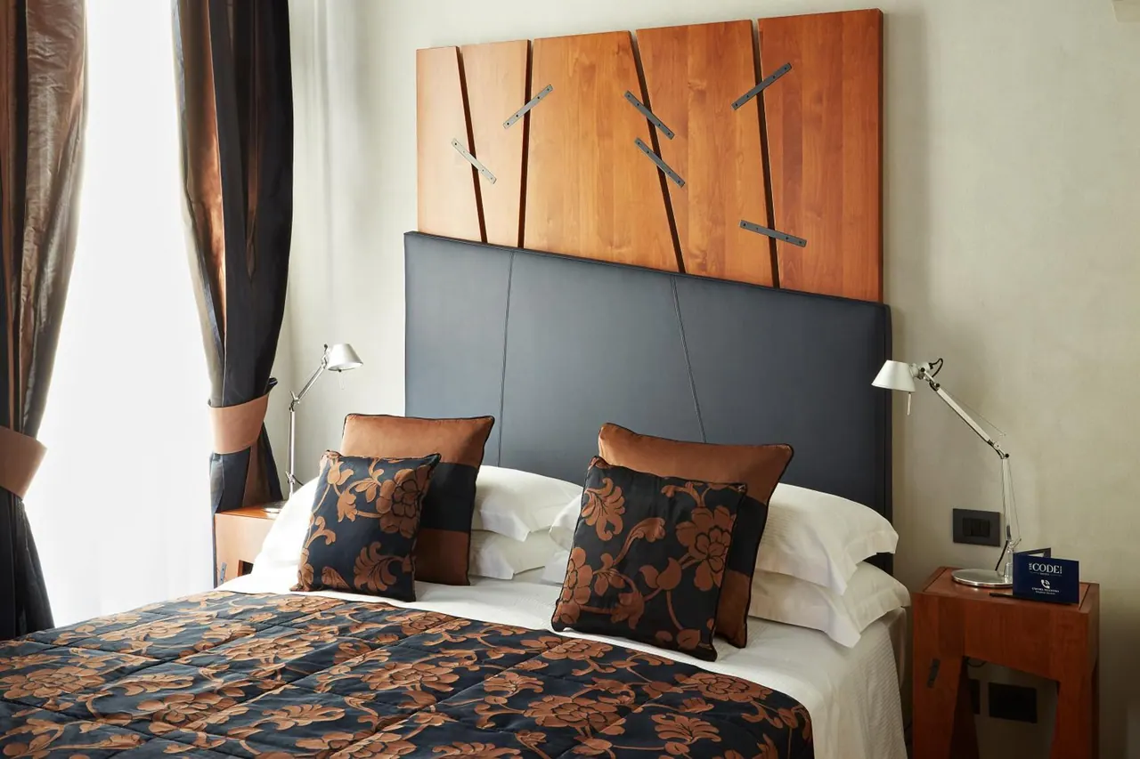 A made double bed with a dark floral throw and decorative pillows sits beneath a wooden headboard panel, flanked by wall-mounted reading lamps and wooden bedside tables.
