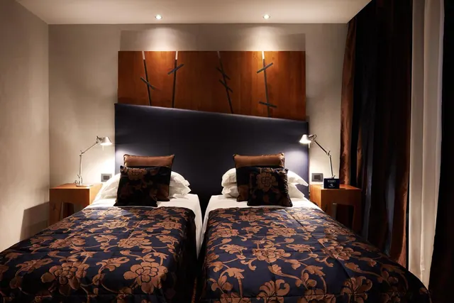 Two single beds with dark floral bedspreads and wooden headboard panels sit side by side in a room lit by bedside lamps and ceiling spotlights.
