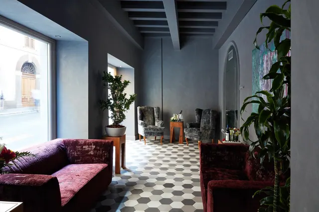 A lounge with burgundy velvet sofas, grey armchairs, a hexagonal tiled floor and a large window onto a street.