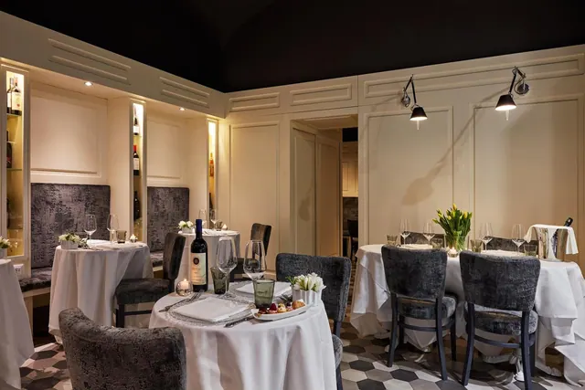 A dimly lit restaurant dining room with white-clothed round tables, grey upholstered chairs, banquette seating and a wine bottle on one table.