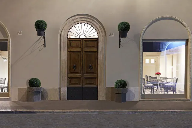 Arched wooden double doors and a large arched window showing a dining room sit in a plastered building facade at night, with topiary planters on the wall and pavement.