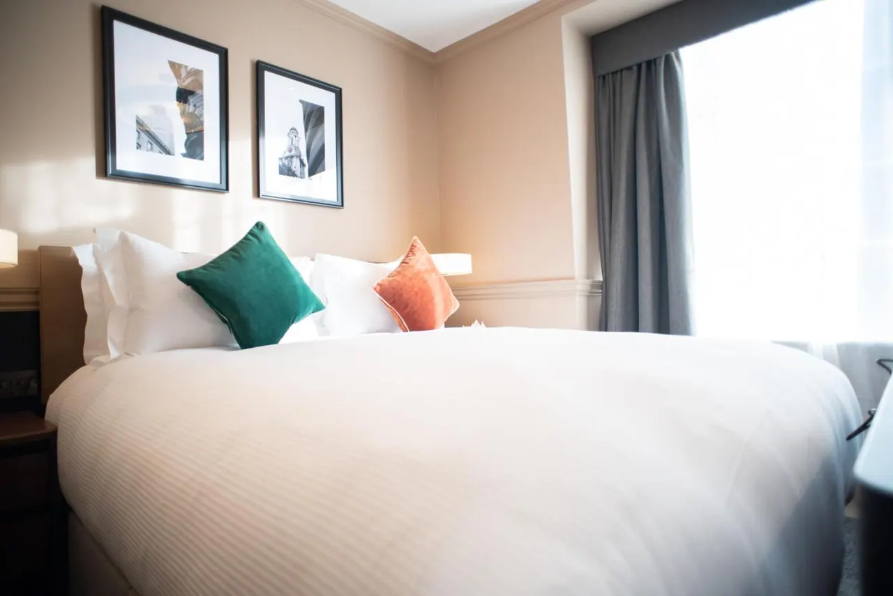 A made-up double bed with white linen and green and orange cushions sits beside a window with grey curtains, with two framed prints on the wall above.