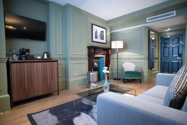 A hotel living area with a grey sofa, a glass coffee table, a fireplace, a wall-mounted television and a wooden sideboard holding a kettle and coffee machine.