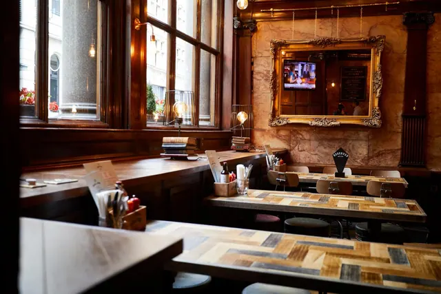 A restaurant dining room with wooden tables and stools, a gold-framed mirror above a marble wall, a wall-mounted television, and windows looking onto a street.