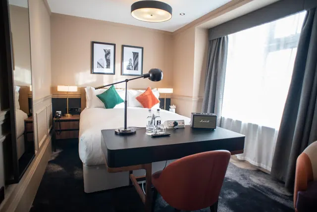 A hotel bedroom with a double bed, a dark desk with a lamp, water bottles and a speaker, and a large window with grey curtains.