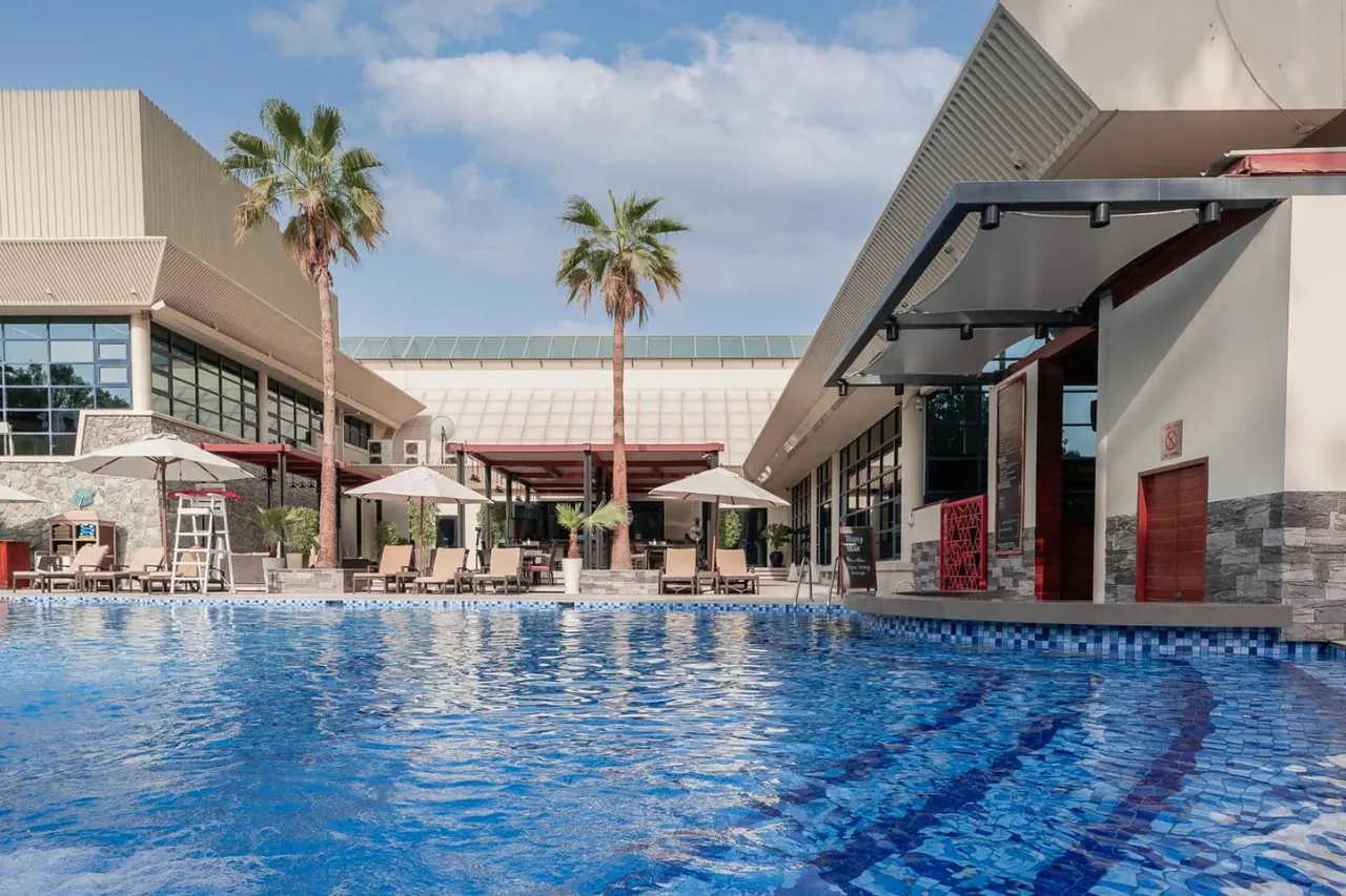 A large outdoor swimming pool with blue mosaic tiles, with sun loungers and white parasols beside it, palm trees and hotel buildings behind.