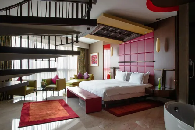 A bedroom suite with a large bed against a pink padded headboard wall, olive green armchairs and a sofa, a loft staircase overhead, and a freestanding bathtub at the right edge.