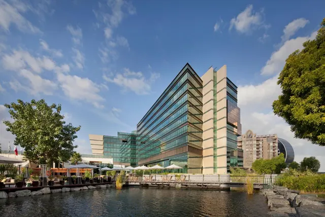 A tall glass-fronted hotel building rises beside a canal, with white umbrellas and sun loungers on a waterside deck and trees on both sides of the frame.