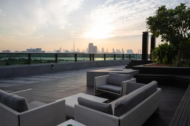 A rooftop terrace with white wicker sofas and a low fire pit table looks out over a city skyline with the Burj Khalifa at sunrise or sunset.