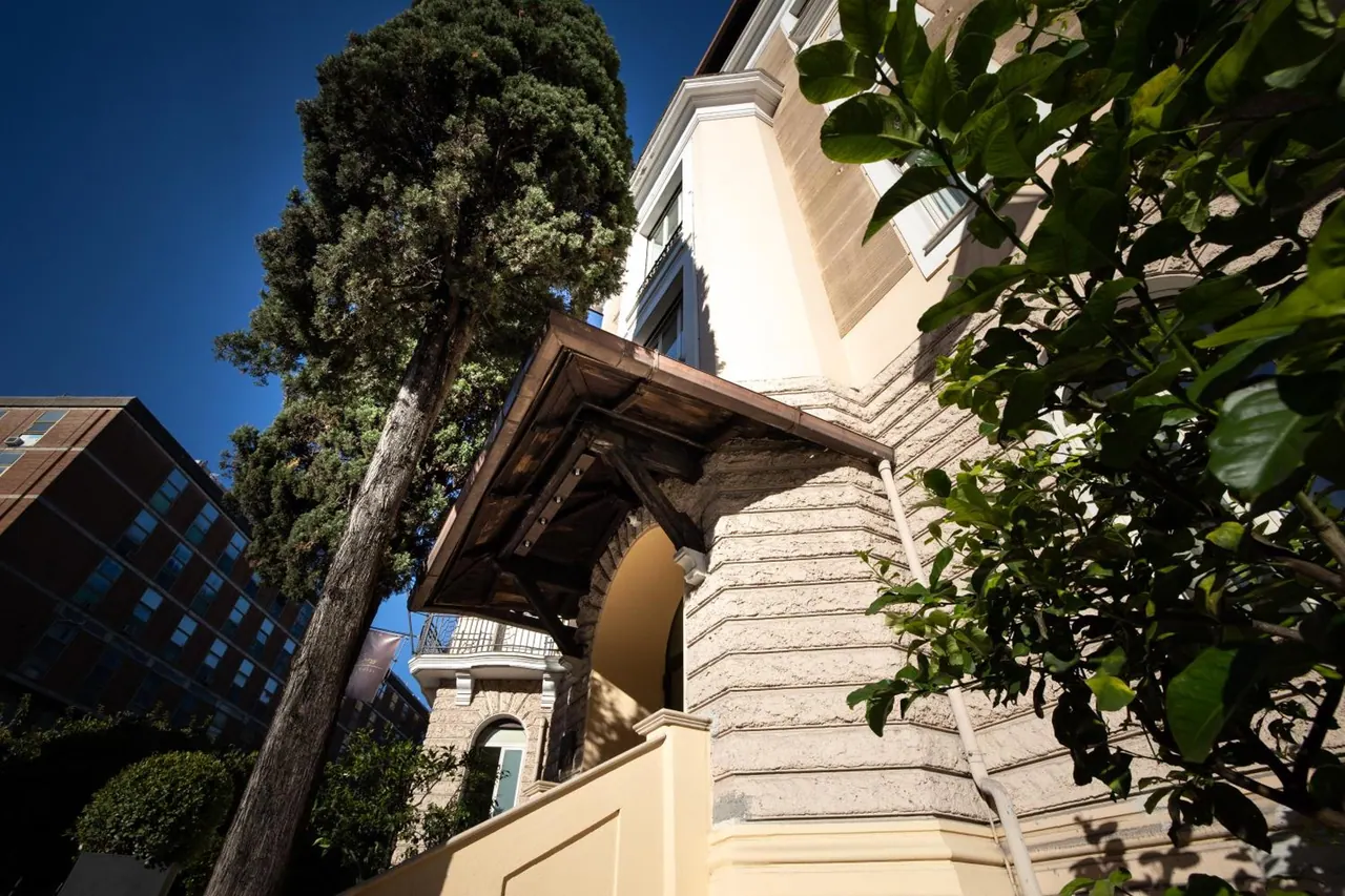 Low-angle view of a cream-coloured building with a wooden canopy over an arched entrance, beside a tall pine tree and a leafy citrus tree under a clear blue sky.