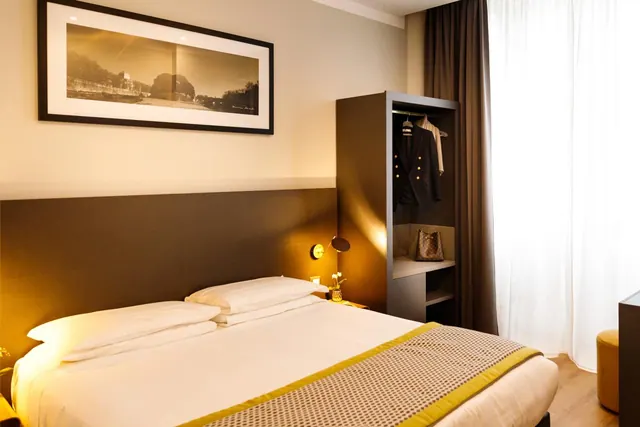 A double bed with white linen and a patterned runner sits in a hotel room beside an open wardrobe with hanging clothes, under a framed sepia print of a city scene, with sheer curtains at the window.