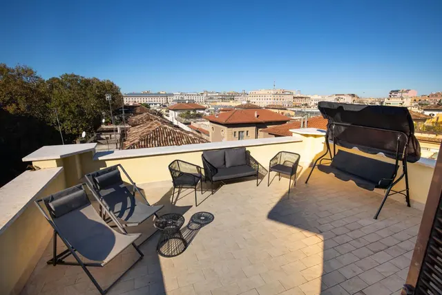 A rooftop terrace with lounge chairs, a swing bench and wicker seating, overlooking terracotta rooftops and buildings under a clear blue sky.