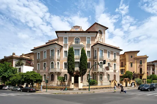 The front of a cream-coloured multi-storey building with a tiled roof and tall cypress trees stands on a street corner, with cars and pedestrians along the road.
