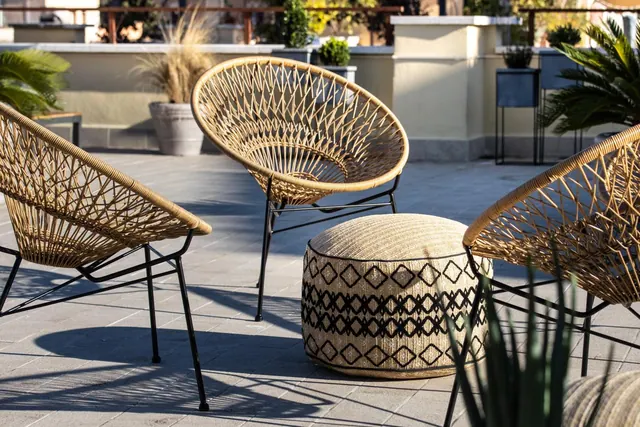 A rooftop terrace with two wicker lounge chairs, a patterned pouf and potted plants along a pale wall.