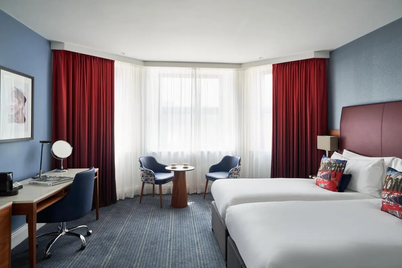 A hotel bedroom with two beds, a desk with a chair, two armchairs beside a round table, and red curtains framing a bay window.