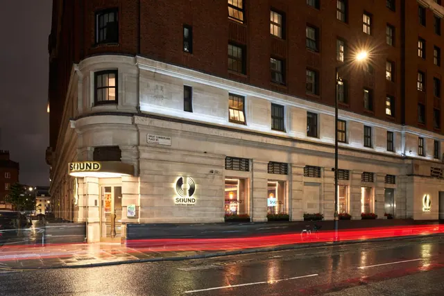 Night exterior of a brick and stone building with illuminated SOUND signage, a street lamp, and red vehicle light trails on a wet road.